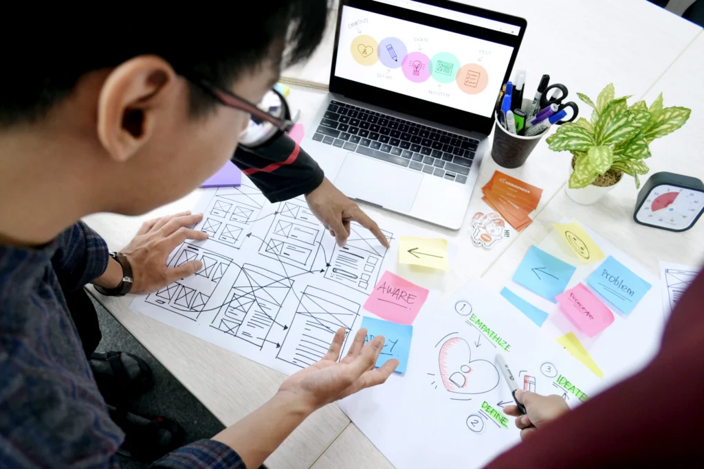 Diseñador organiza bocetos y notas adhesivas junto a una computadora portátil